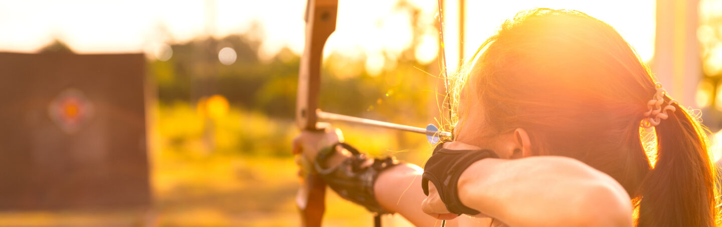 Young female archer aiming at target in field at sunset time