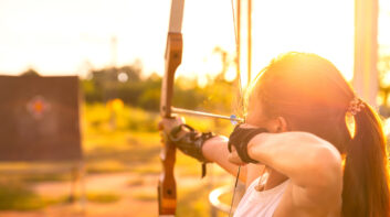 Young female archer aiming at target in field at sunset time