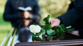 Fresh white rose being put by woman in mourning attire on closed coffin lid